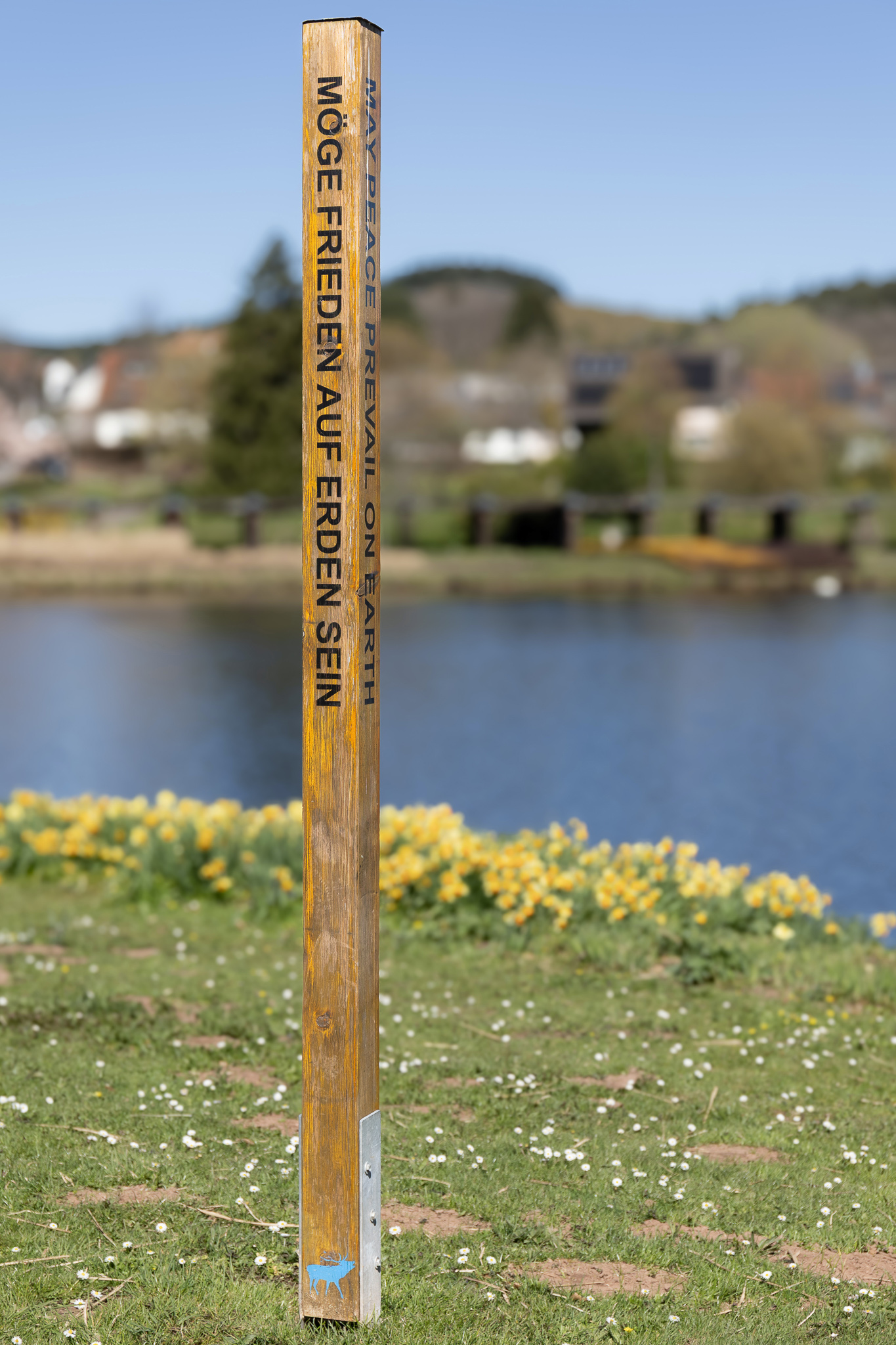 Holzpfosten mit der Aufschrift 'Möge Frieden auf Erden sein' im Kurpark Weiskirchen vor unscharfem Hintergrund